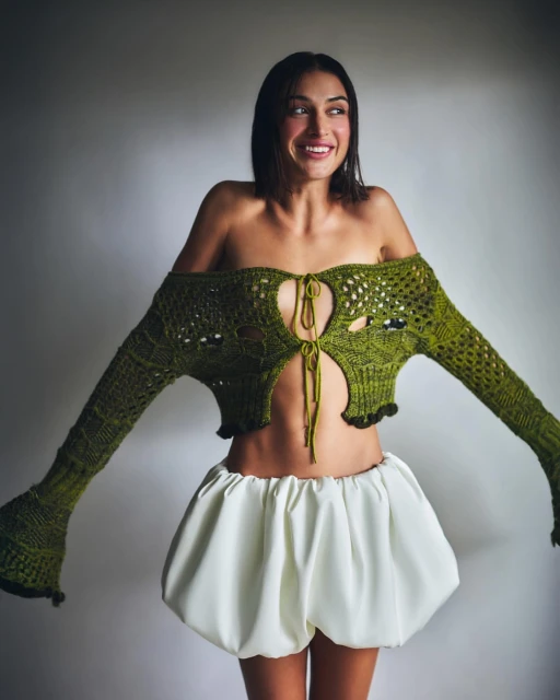 Smiling woman in a green crochet cardigan and white bloomer shorts poses against a neutral background in a fashion portrait.