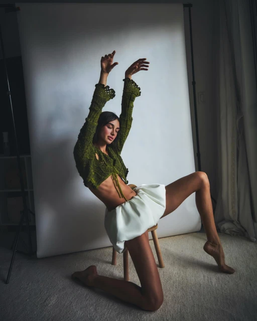 A young woman in an olive knit top and white skirt poses barefoot on a stool in a photo studio against a white backdrop.