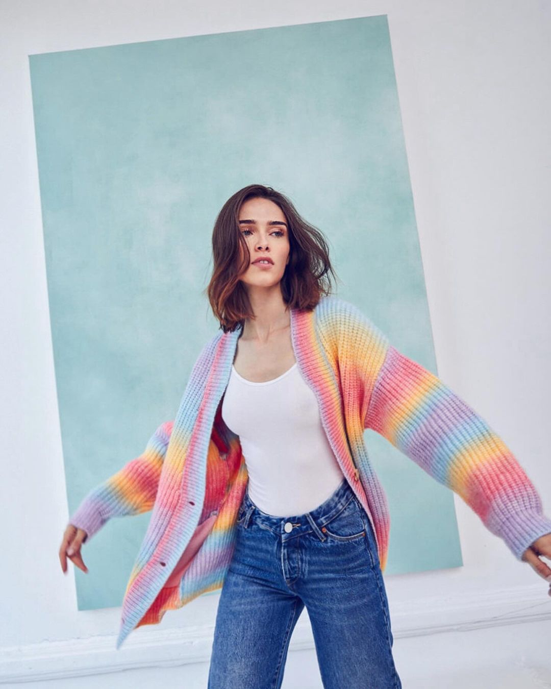 Woman wearing a colorful striped cardigan, white top, and jeans poses against a blue backdrop in a fashion studio shot.