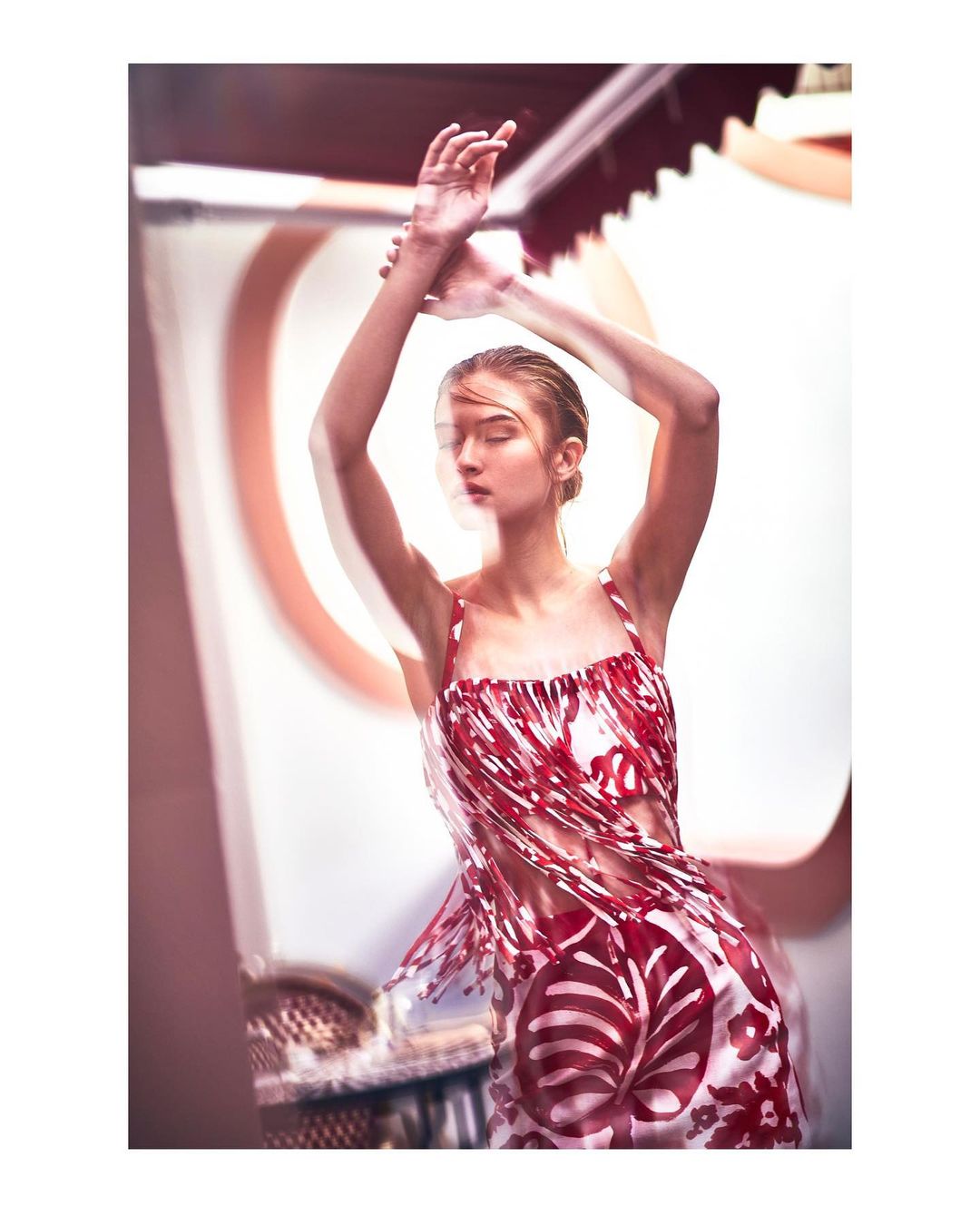 Woman poses in patterned dress with raised arms and blurred motion effect in bright indoor setting with architectural details.