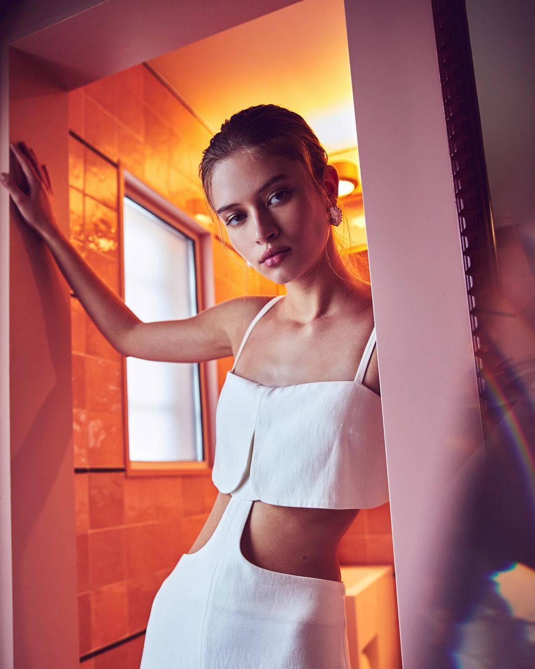 Woman with light skin and hair poses in a white dress with cutout near a doorway and bathroom in the background with peach tile.
