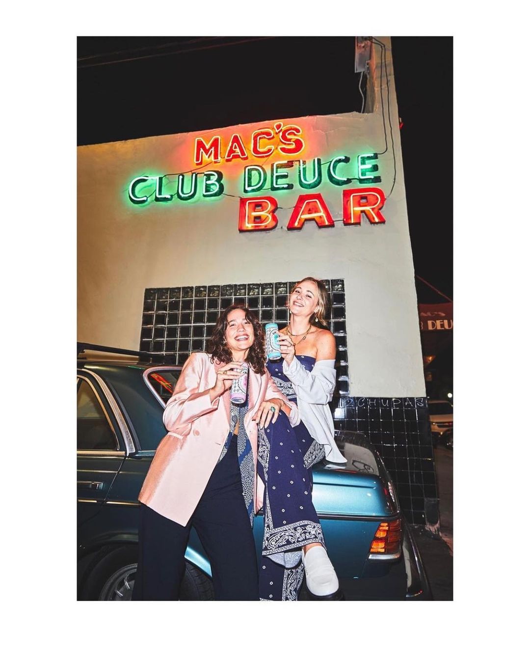 Two women pose with drinks on a car under the neon lights of Mac's Club Deuce Bar, enjoying an evening out in an urban setting.