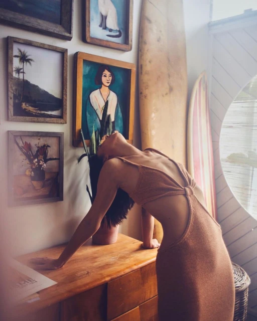 Indoor shot of a woman in a brown dress leaning on a wooden dresser, surrounded by framed artwork and surfboards, with natural light.