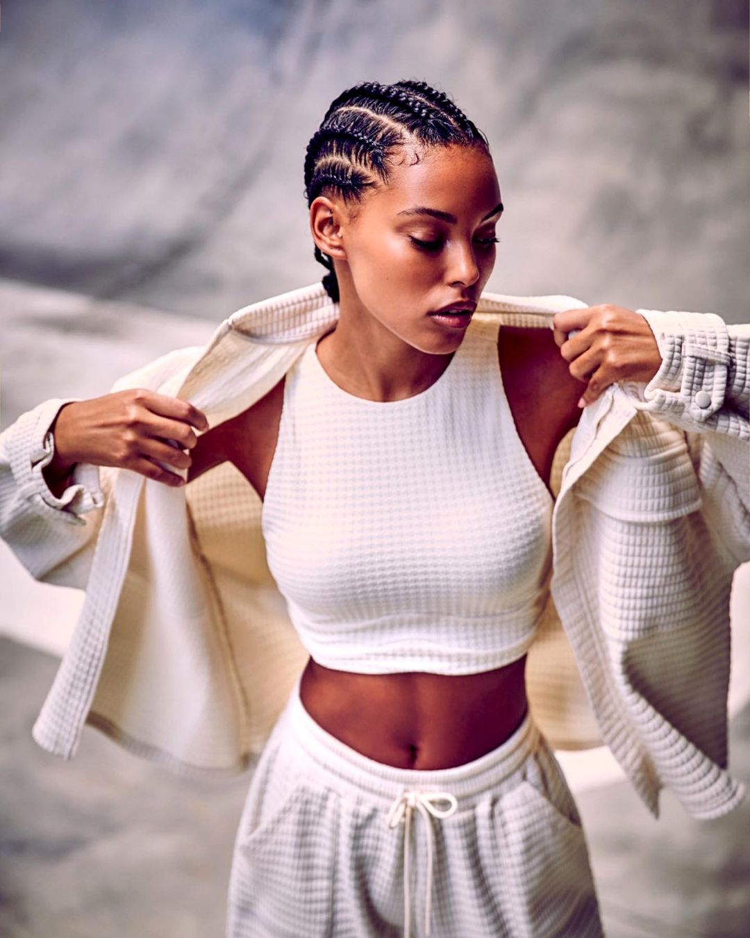 Woman in a matching athletic crop top, jacket, and sweatpants poses in front of a grey mottled background.