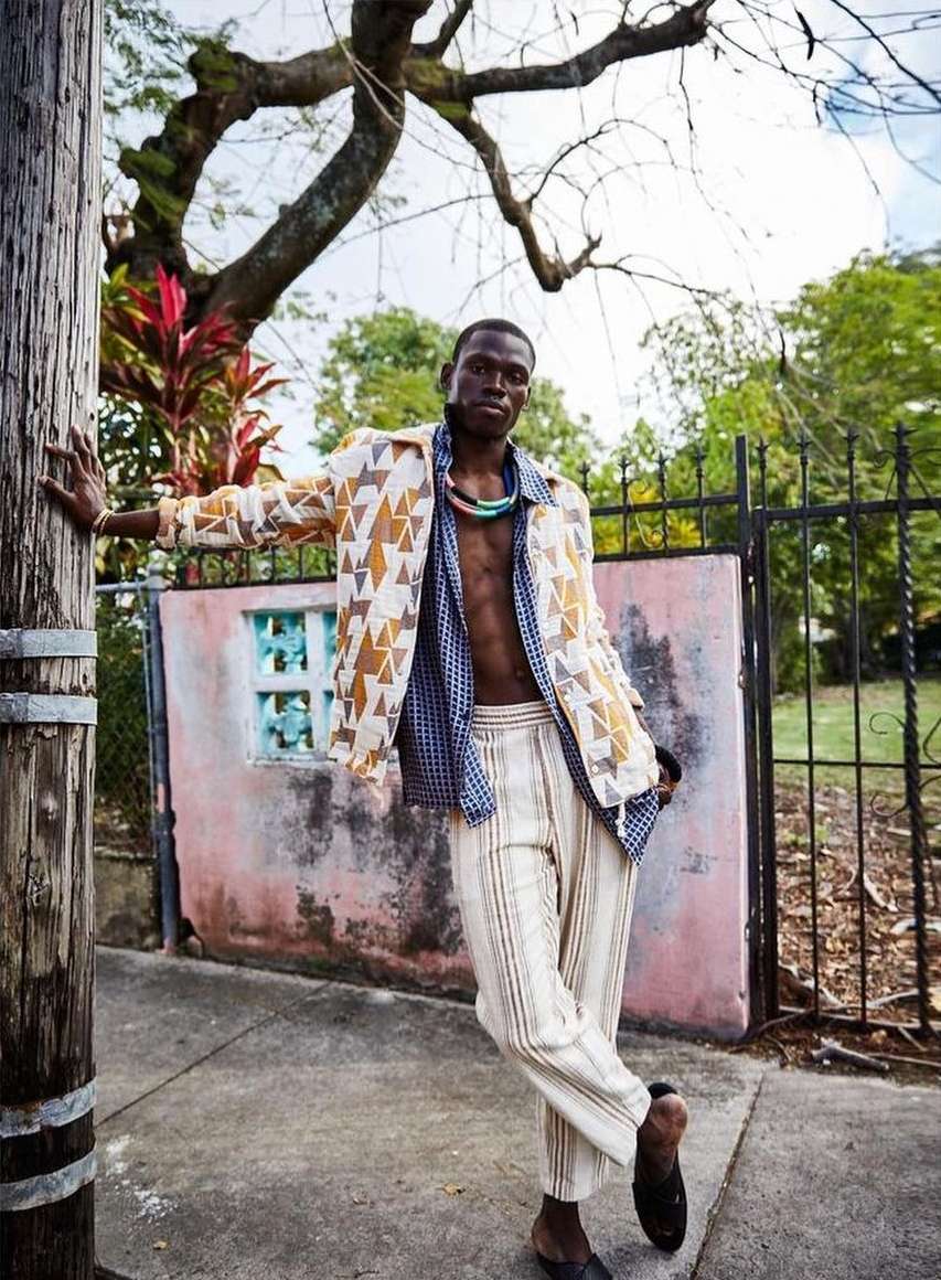 A Black male model leans against a wooden pole outdoors, wearing an open blue shirt, patterned jacket, striped pants, and necklace.