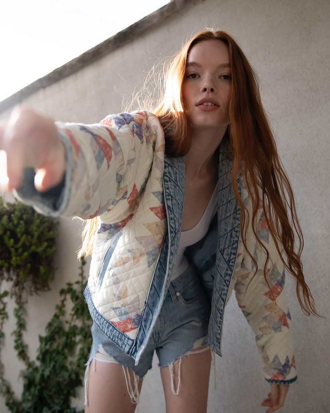 A young woman with long red hair poses in a denim-trimmed jacket with quilted patchwork, pointing one arm forward in a natural outdoor setting.
