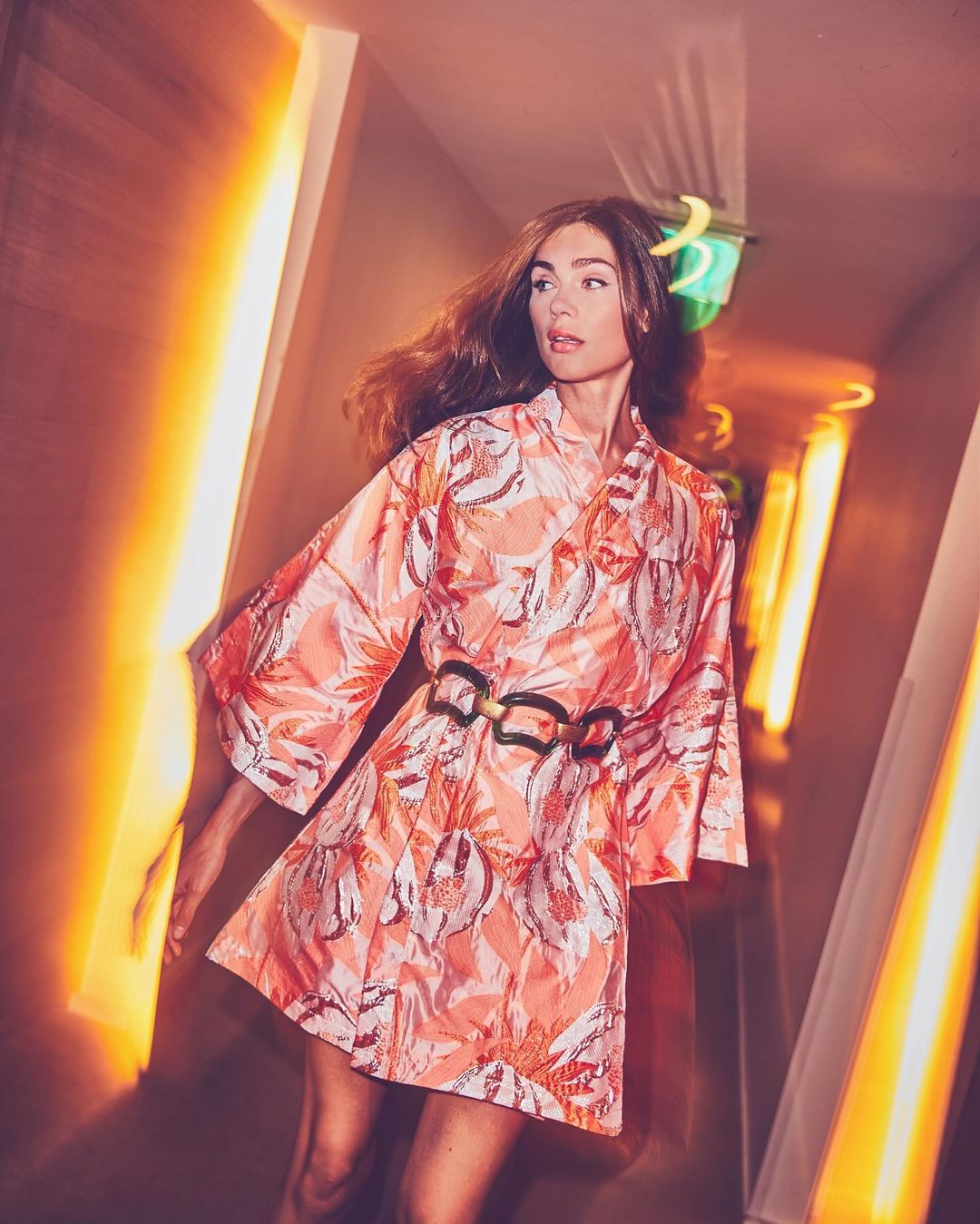 Attractive adult female wearing patterned dress with belt poses in a corridor with bright geometric orange and white lighting.