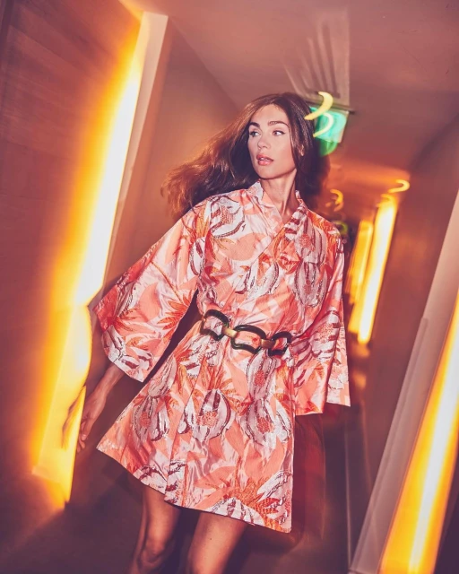 Attractive adult female wearing patterned dress with belt poses in a corridor with bright geometric orange and white lighting.