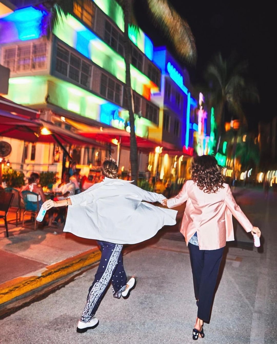 Two women stroll hand-in-hand at night along a city street with brightly lit buildings and palm trees in the background.