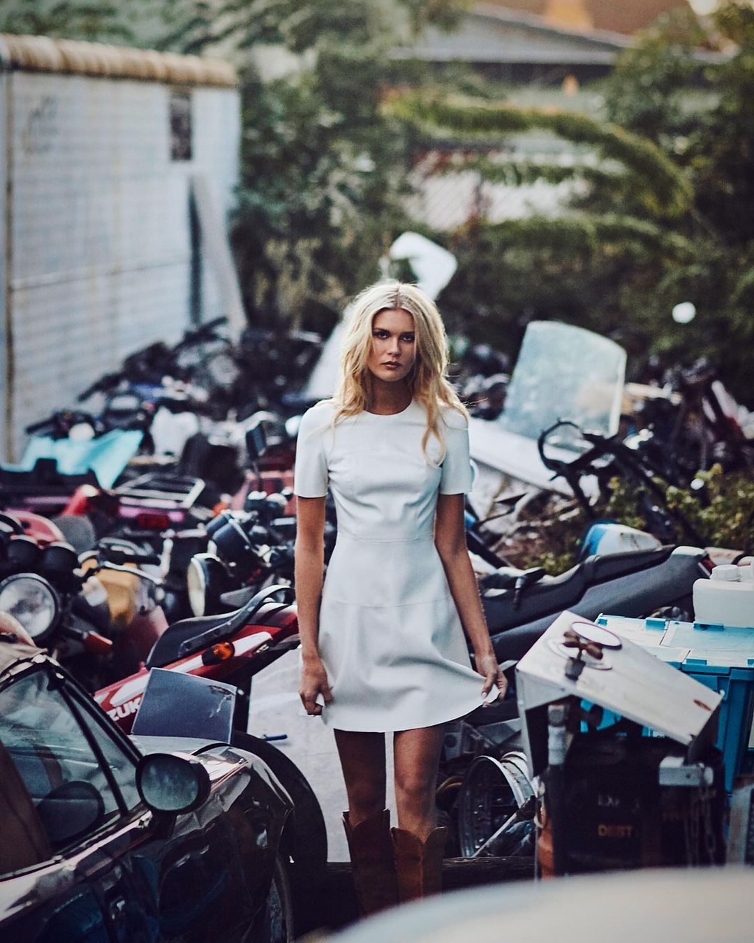 Blonde woman in white dress and boots stands in a cluttered outdoor environment with vintage cars and auto parts.