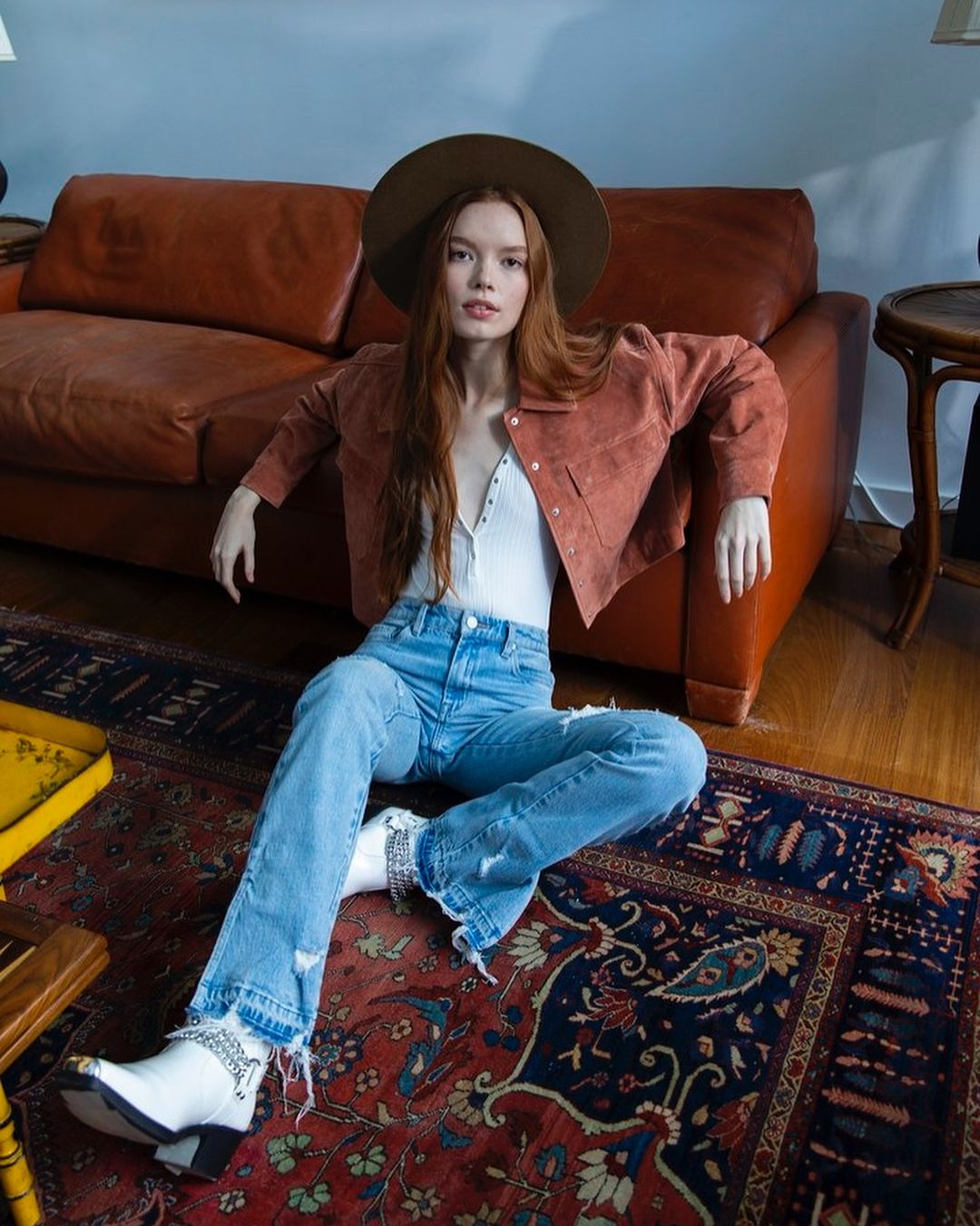 Stylish woman with long auburn hair wears a hat, jacket, jeans, and boots while posing indoors on a patterned rug near furniture.