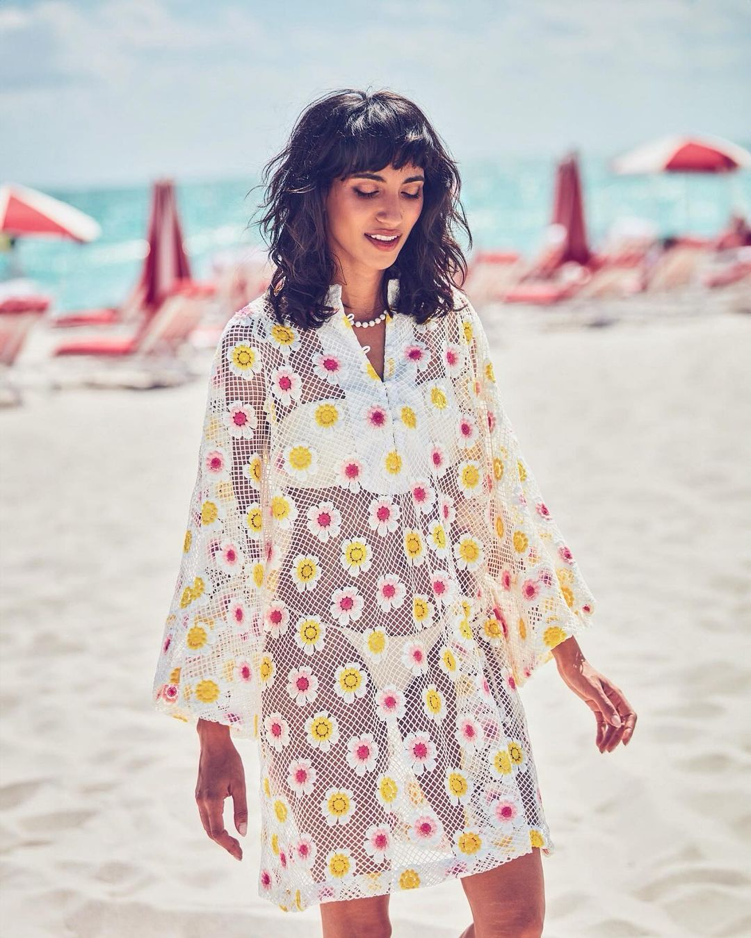 Adult female on a sunny beach wearing a white floral-patterned cover-up, with the ocean and beach umbrellas visible in the background.