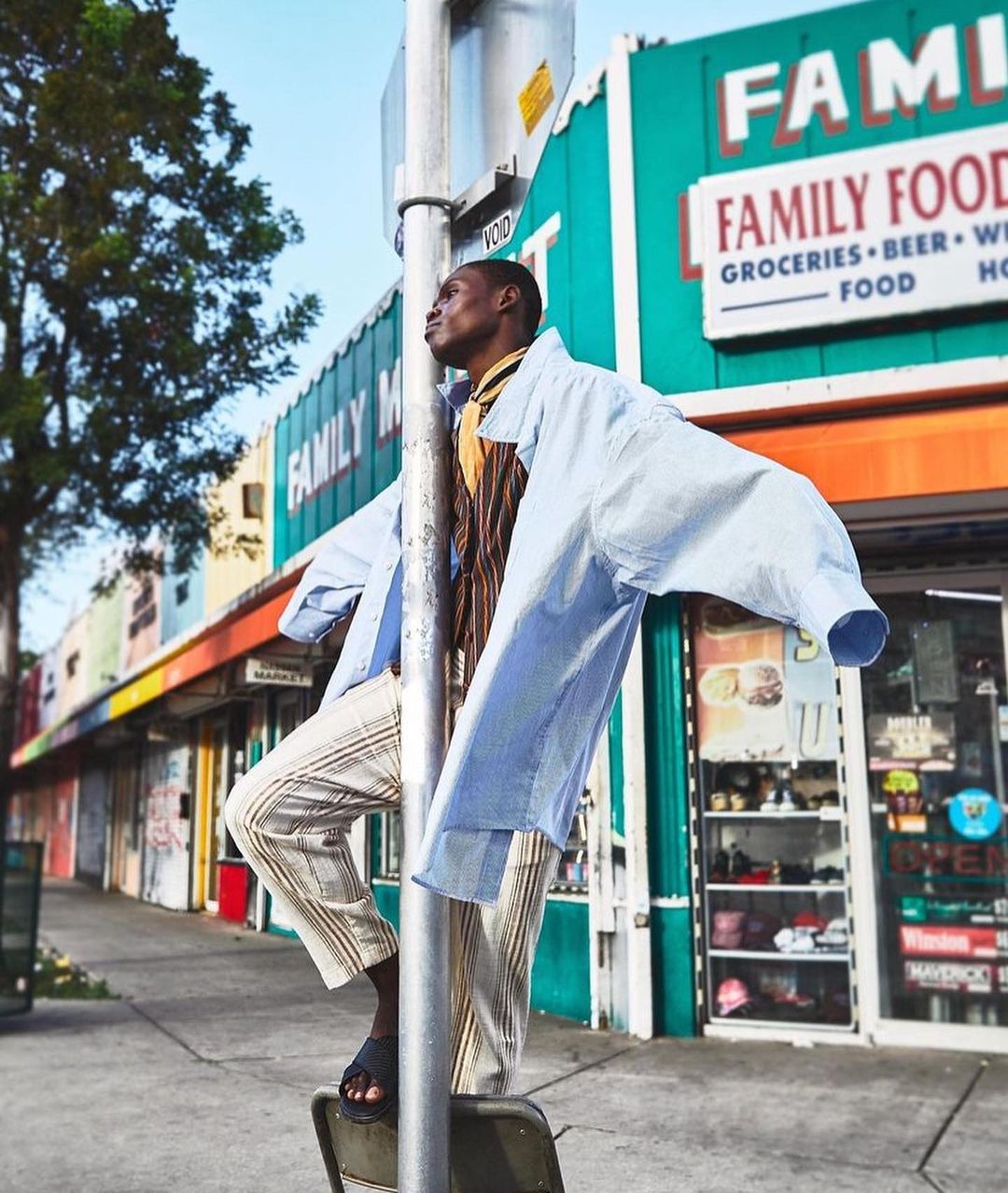 Stylish man in casual attire poses by a pole next to a Family Food grocery store on a sunny urban street. Fashion editorial shot.