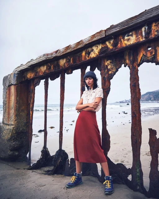 Woman posing on a beach in skirt and beanie, leaning against a weathered barrier, ocean background, cloudy day, casual fashion aesthetic.