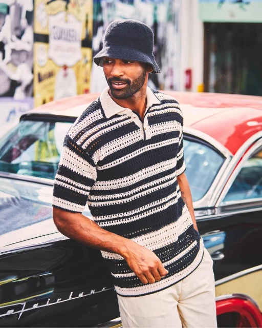 Adult male in striped knit shirt and bucket hat leans on vintage car in urban setting with colorful graffiti art in the background.