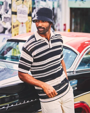 Adult male in striped knit shirt and bucket hat leans on vintage car in urban setting with colorful graffiti art in the background.