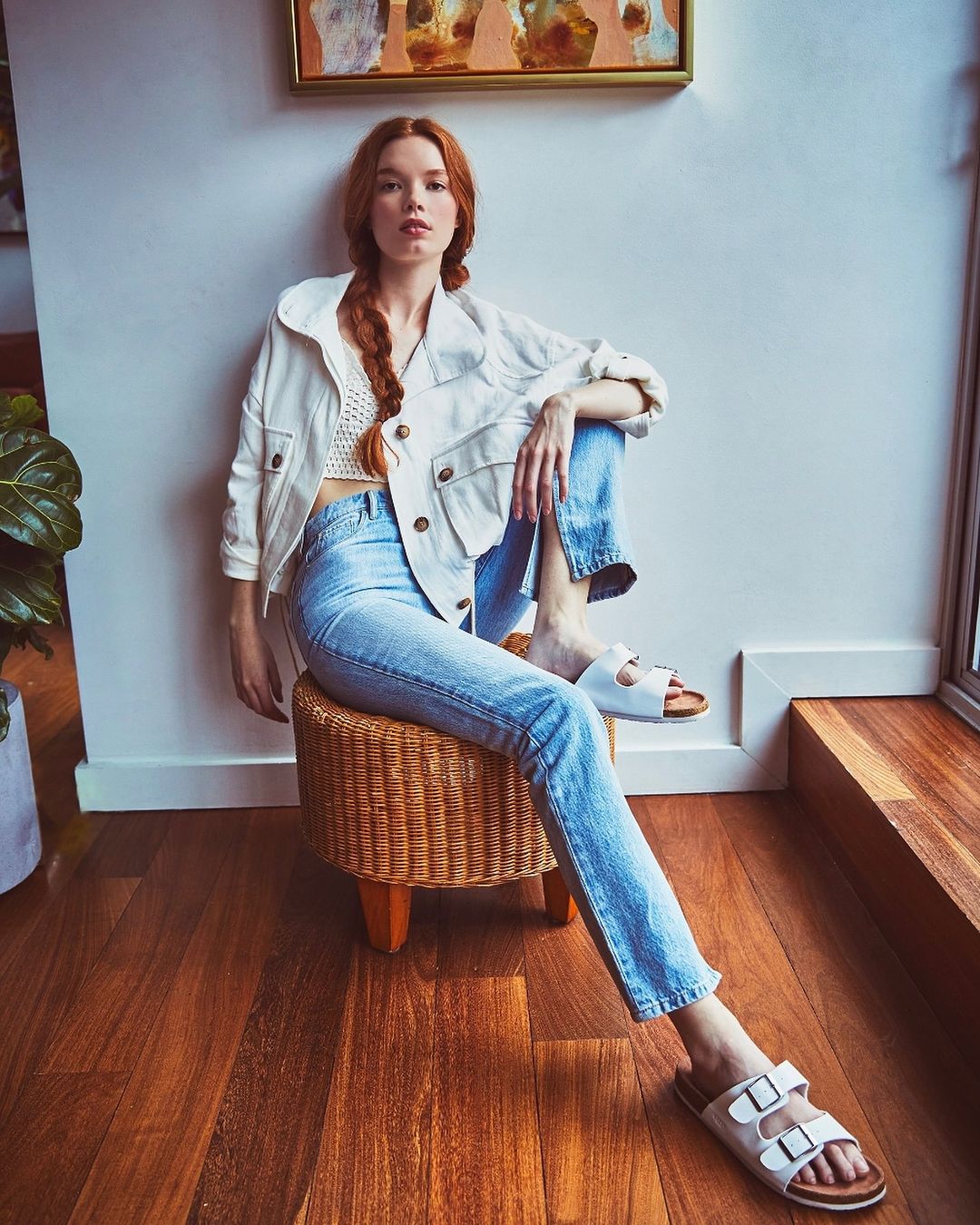 A red-haired woman sits on a wicker stool indoors. She wears a white jacket, blue jeans, and white sandals near a window and a green plant.