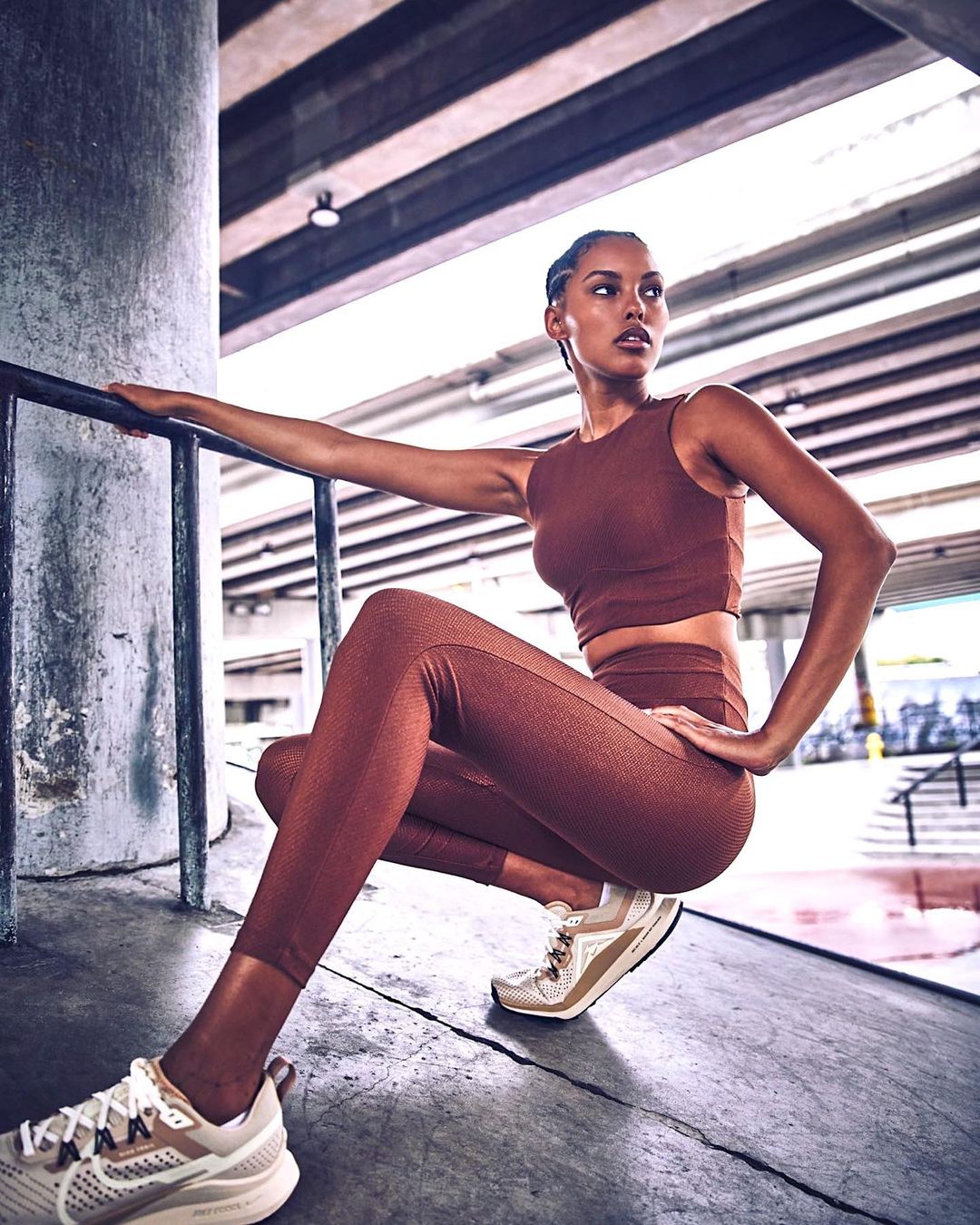 Athletic woman in brown workout clothes crouching on concrete in an urban setting, wearing running shoes and posing near a metal railing.