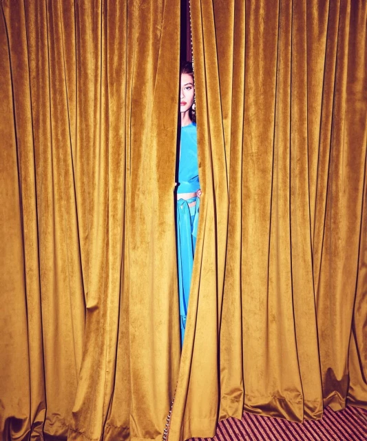 A woman with fair skin in a blue dress peeks out from behind golden velvet curtains in an indoor setting with striped flooring.