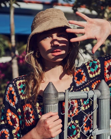 Model wearing a tan bucket hat and colorful patterned crochet top posing behind a chain-link fence outdoors in New York.