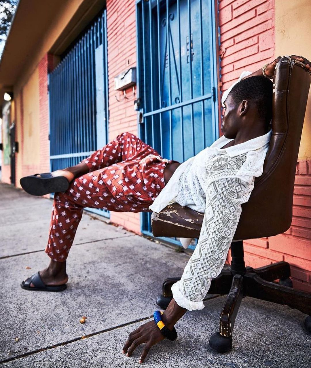 Young Black male lounging in an office chair on a sidewalk in front of a brick building wearing a patterned shirt and pants and sandals.