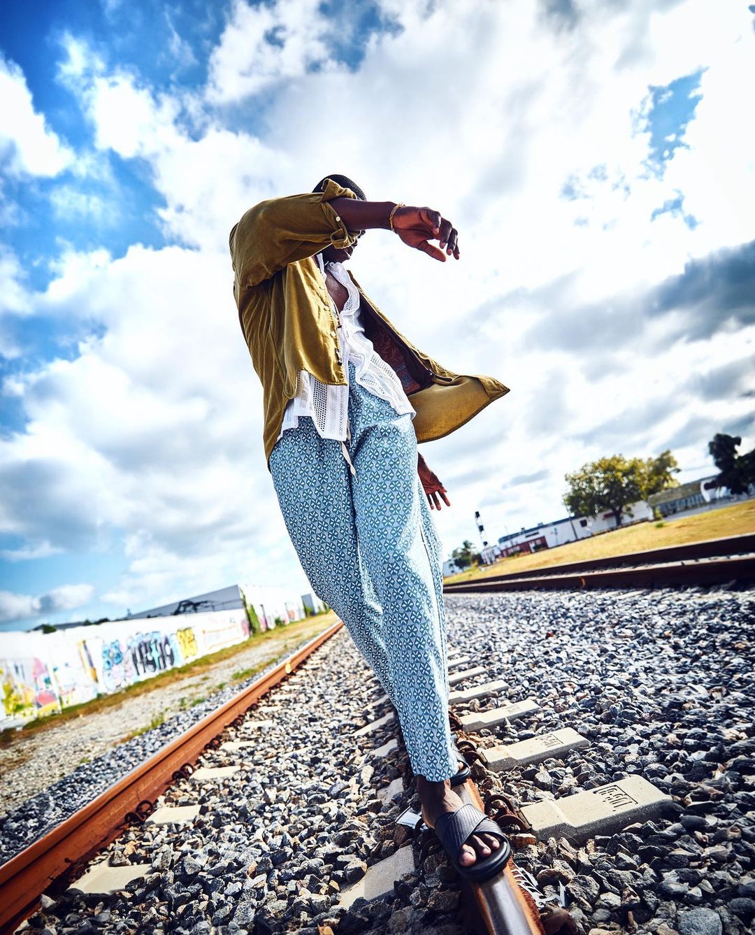 Adult male balances on railroad track outdoors against a cloudy sky, wearing a yellow shirt, white top, and patterned blue pants.