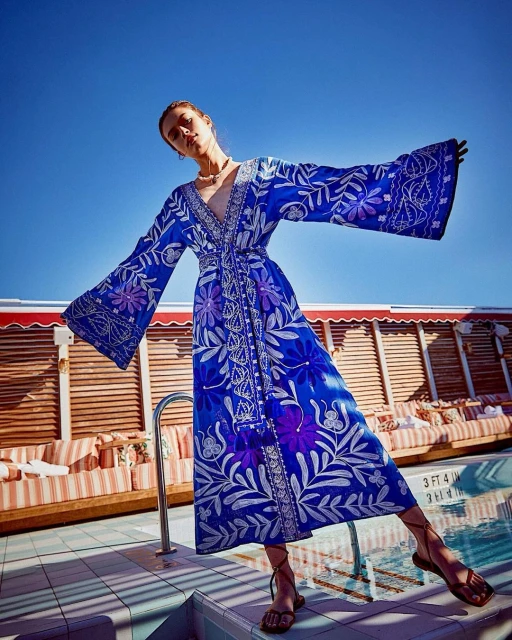 Woman in blue floral robe poses by pool with lounge chairs under bright sky, fashion photography, travel, outdoor setting.