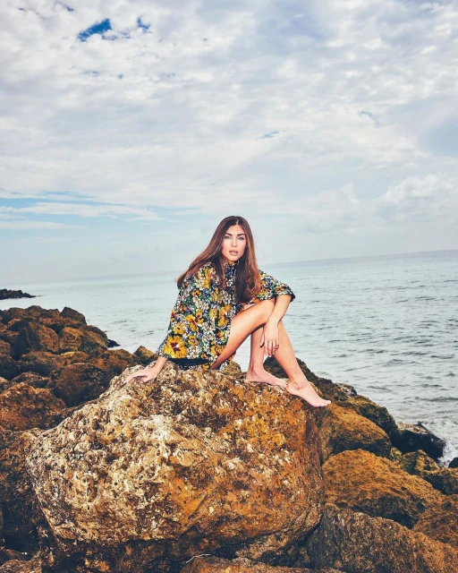 Woman in floral top sits barefoot on a rock near the ocean under a cloudy sky in a medium shot.