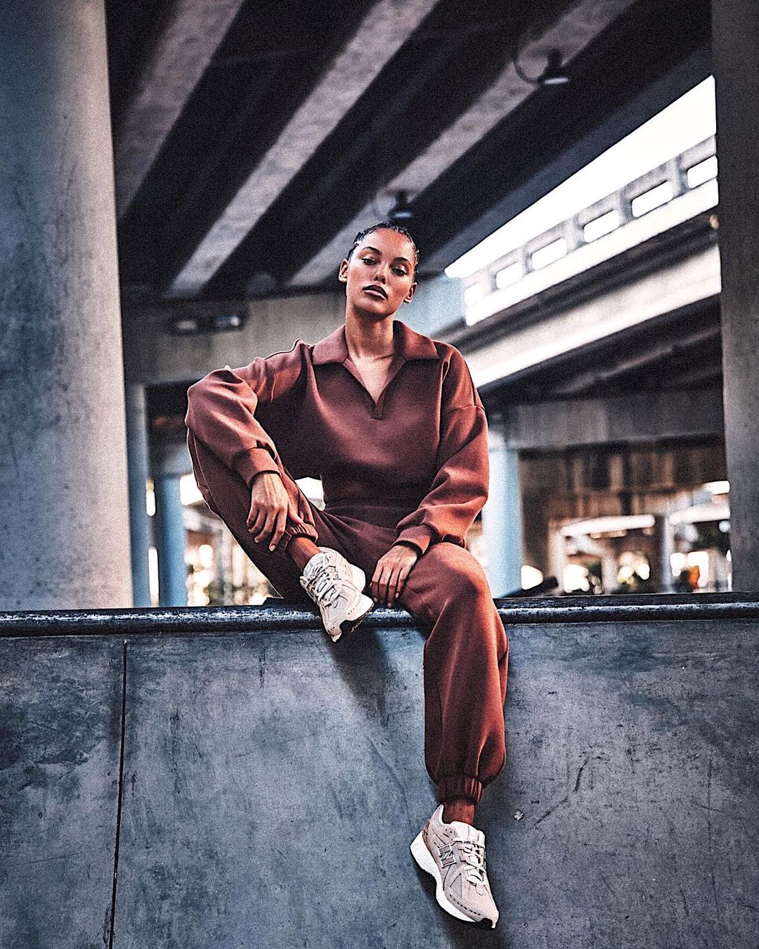 Woman in brown sweatsuit sits on concrete ledge in an urban setting with geometric background of layered highway overpasses, wearing sneakers.