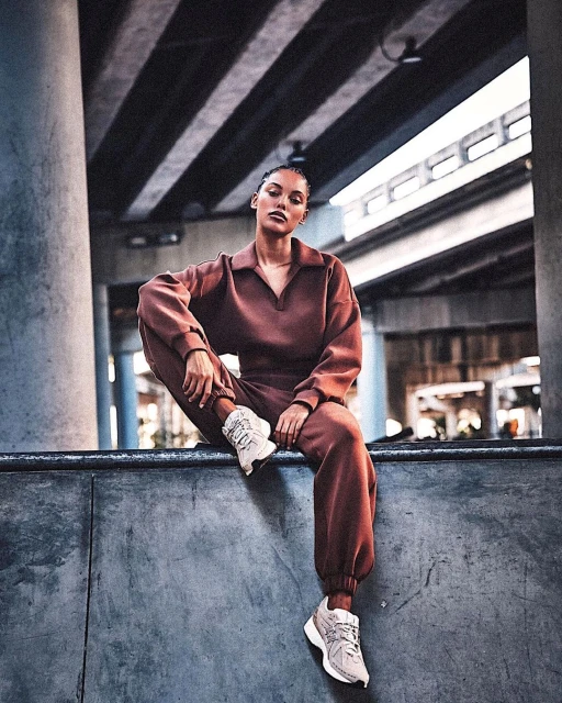 Woman in brown sweatsuit sits on concrete ledge in an urban setting with geometric background of layered highway overpasses, wearing sneakers.
