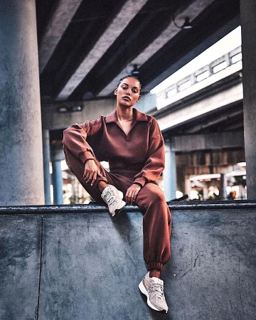 Woman in brown sweatsuit sits on concrete ledge in an urban setting with geometric background of layered highway overpasses, wearing sneakers.