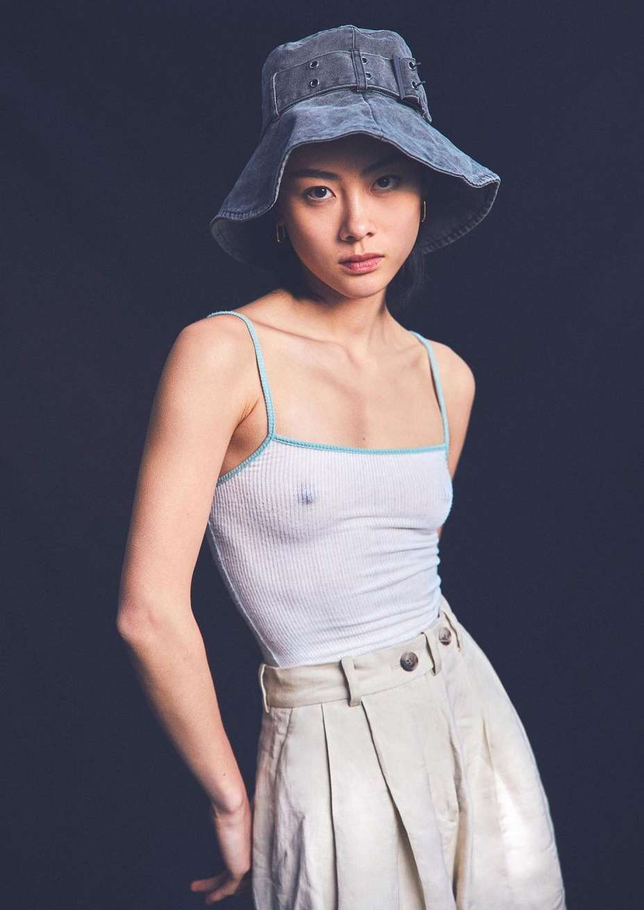 Stylish woman in a white tank top, khaki pleated trousers, and a wide-brimmed denim bucket hat poses in a studio with dark background.