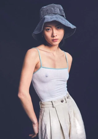 Stylish woman in a white tank top, khaki pleated trousers, and a wide-brimmed denim bucket hat poses in a studio with dark background.