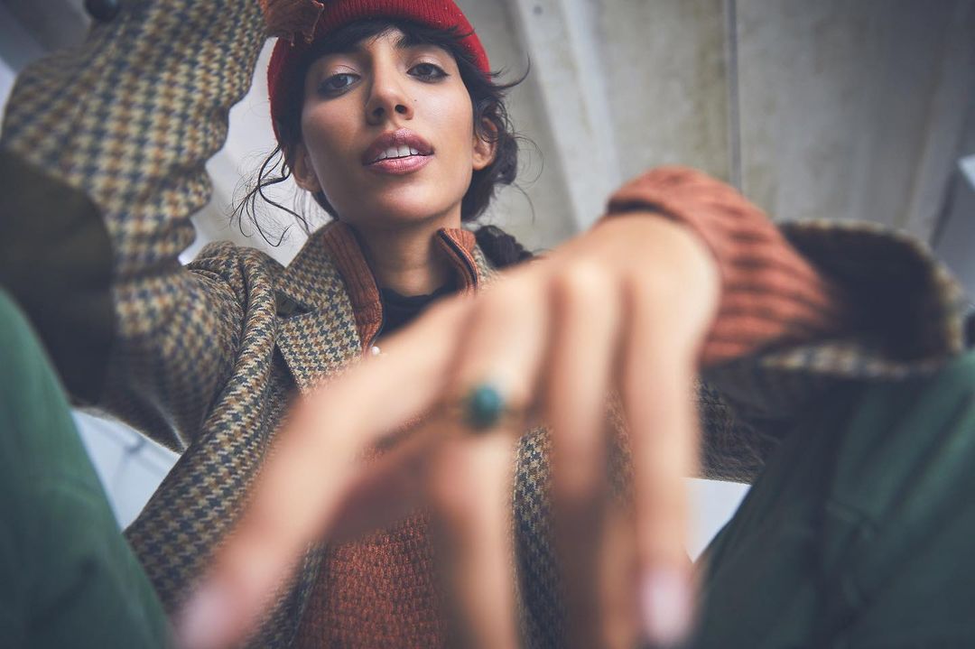 Stylish woman in a red beanie, houndstooth coat, and brown sweater gazes downward with an outstretched hand in an urban setting.