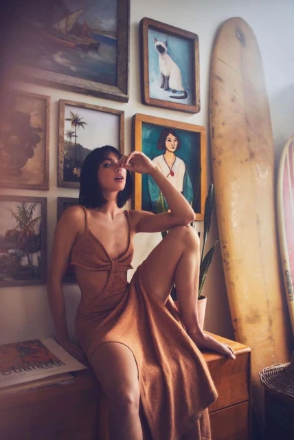 Brunette woman in cut-out dress sits on wooden cabinet with framed artwork, houseplants, and surfboards in the background. Interior shot.
