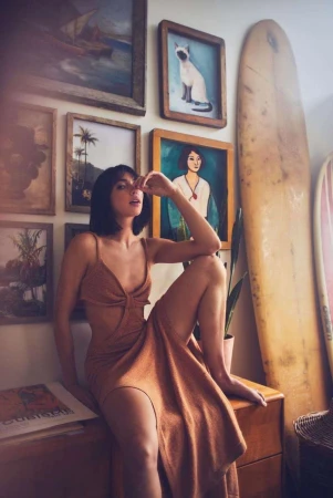 Brunette woman in cut-out dress sits on wooden cabinet with framed artwork, houseplants, and surfboards in the background. Interior shot.