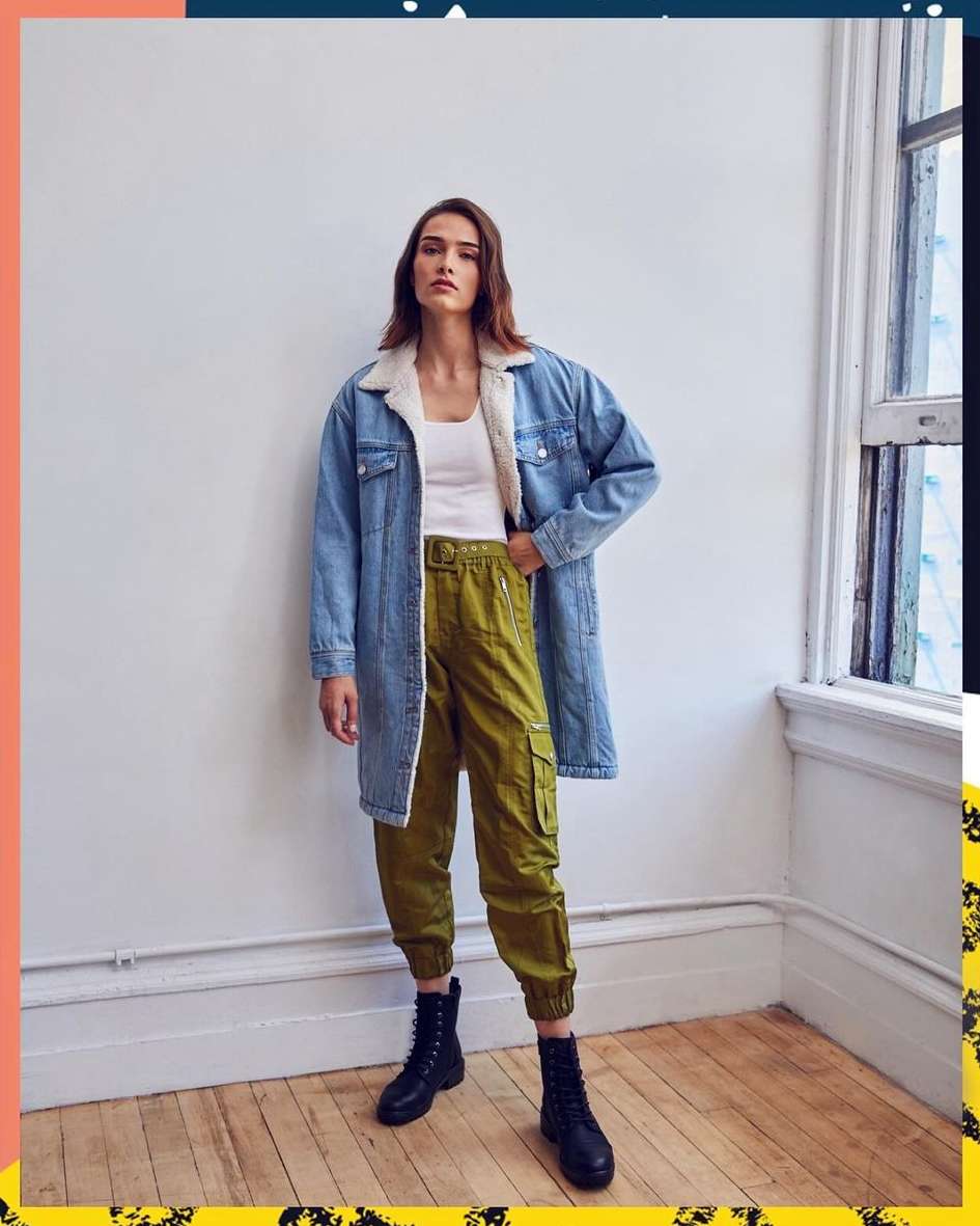 Woman in denim jacket, white top, olive cargo pants, and black boots stands indoors against a white wall with natural window light.