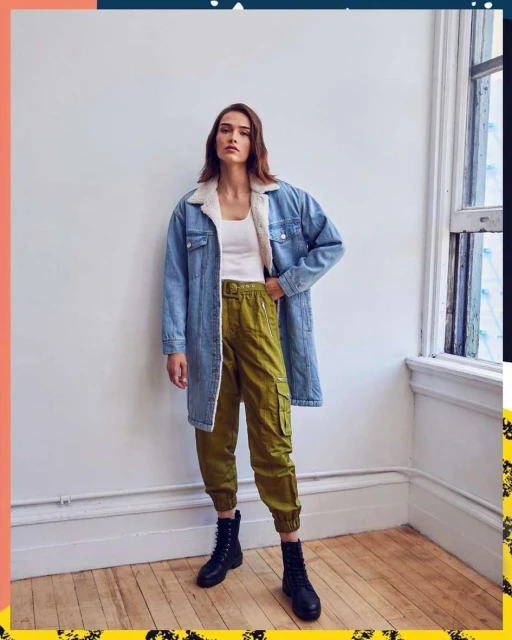 Woman in denim jacket, white top, olive cargo pants, and black boots stands indoors against a white wall with natural window light.