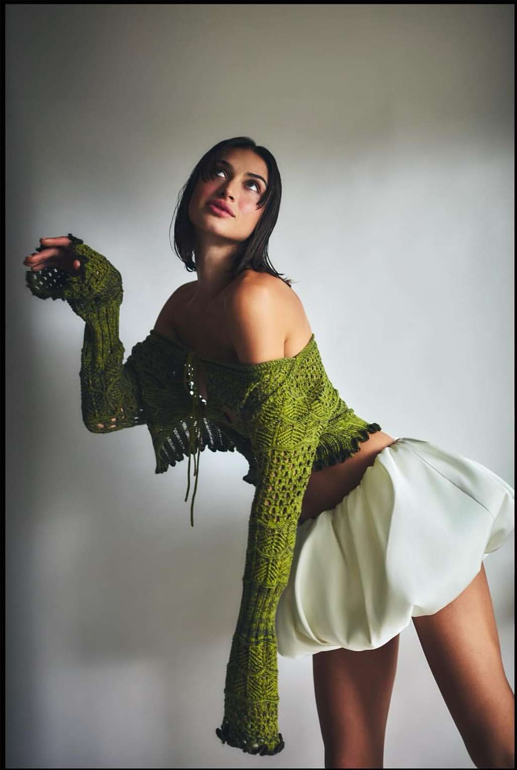 Young woman poses in a green crochet top and white skirt against a grey background, looking up and to the left in a studio setting.