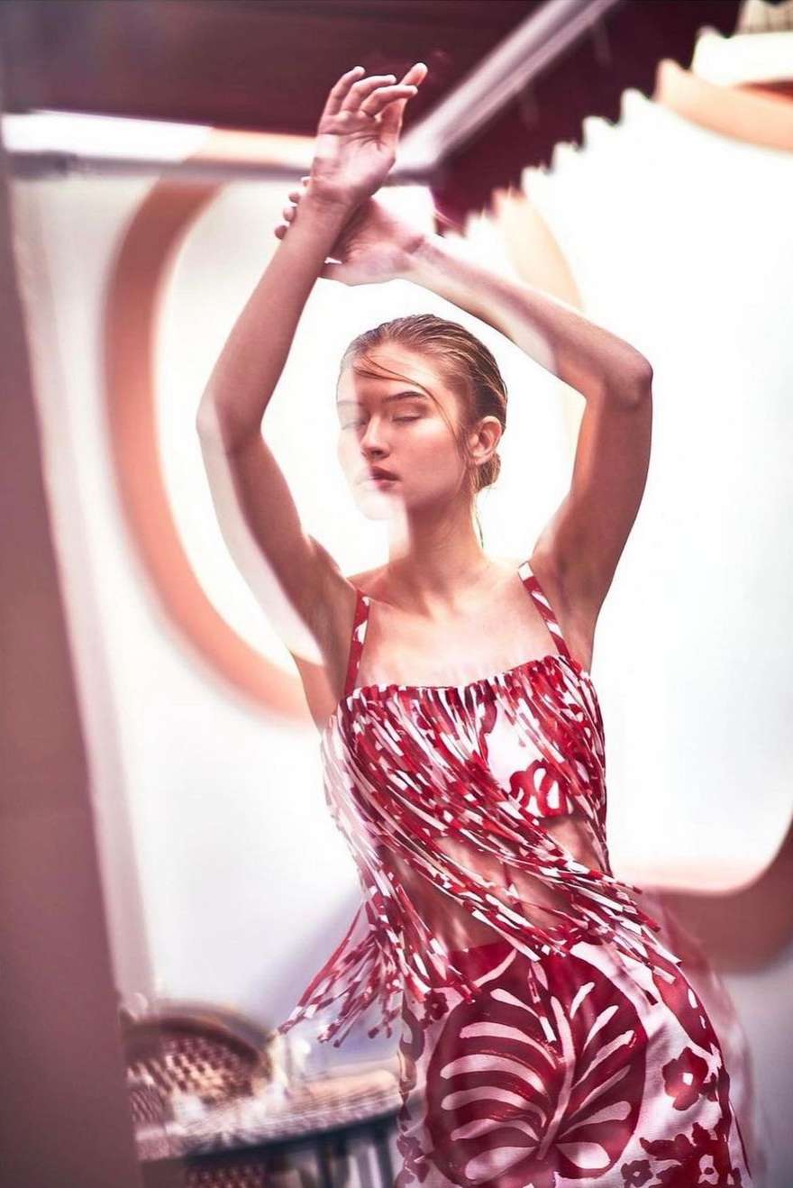 Fashion editorial shot of a young woman wearing a red-and-white floral dress, with arms raised in a light-filled, patterned indoor setting.
