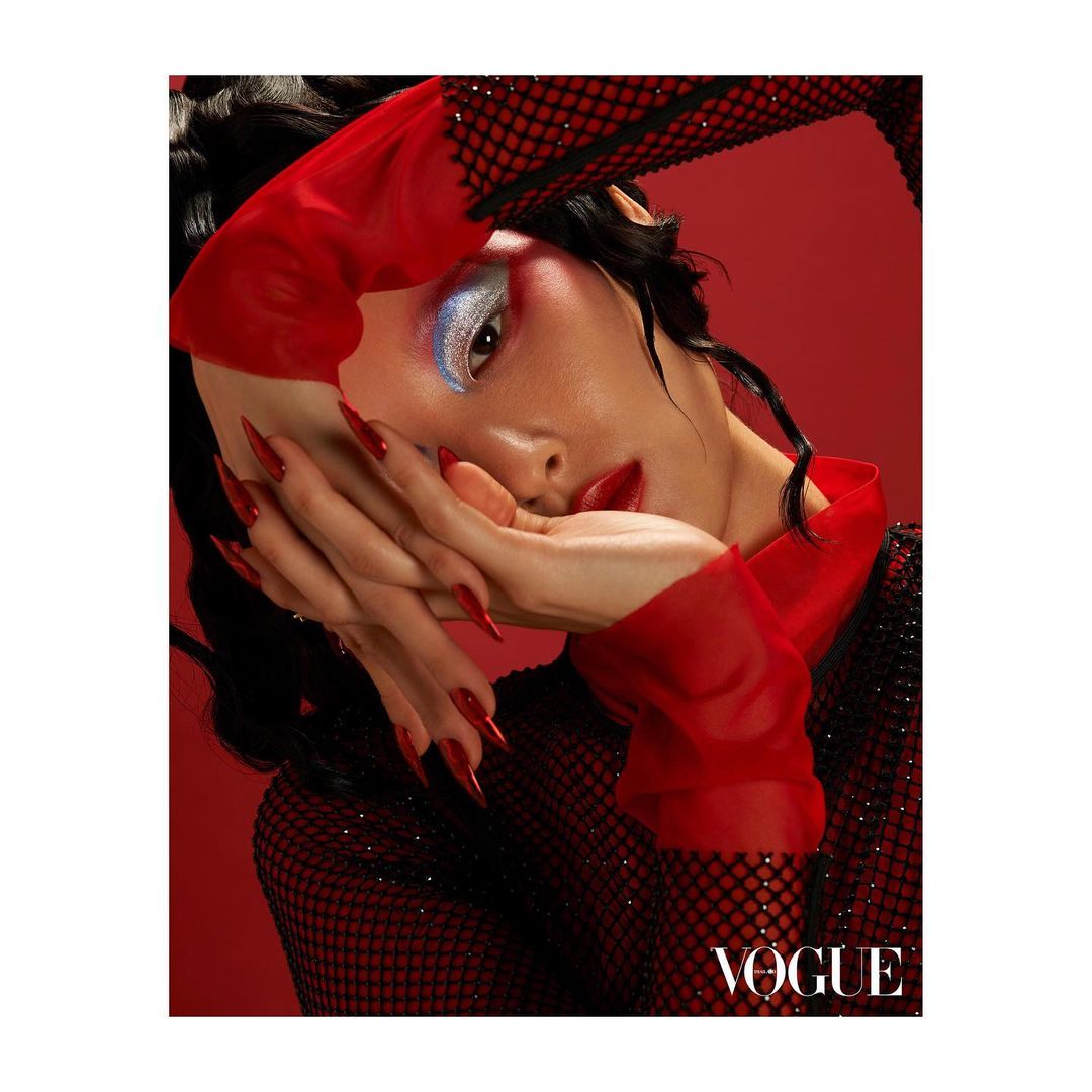 Eye-catching editorial portrait of a woman with red lips, exaggerated makeup, and long red nails, posing in a black fishnet top with a red background.
