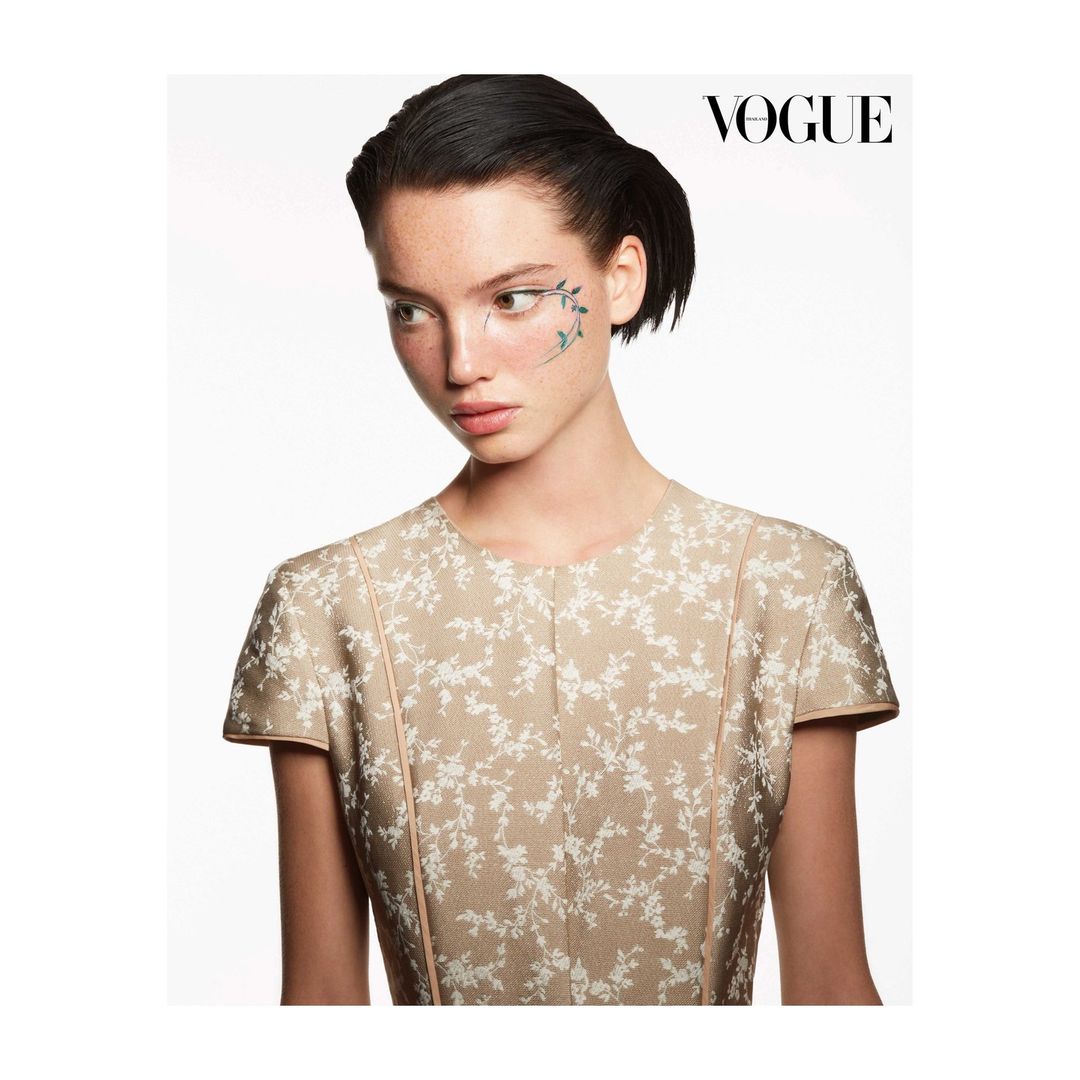 Close-up portrait of a young adult woman in a floral dress with decorative vines painted on her face, Vogue magazine logo visible.