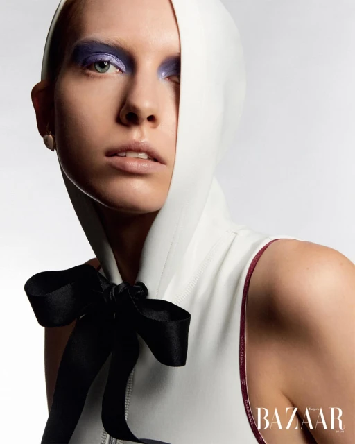 Close-up of a fair-skinned woman with purple eyeshadow, wearing a white hooded garment with a black ribbon bow, and BAZAAR logo.