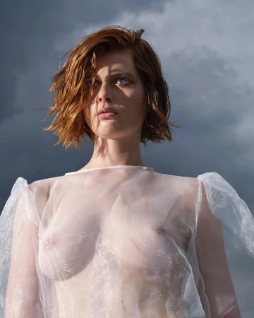 Portrait of a young adult female model with wet red hair and pale skin, wearing a sheer blouse against a cloudy sky background.