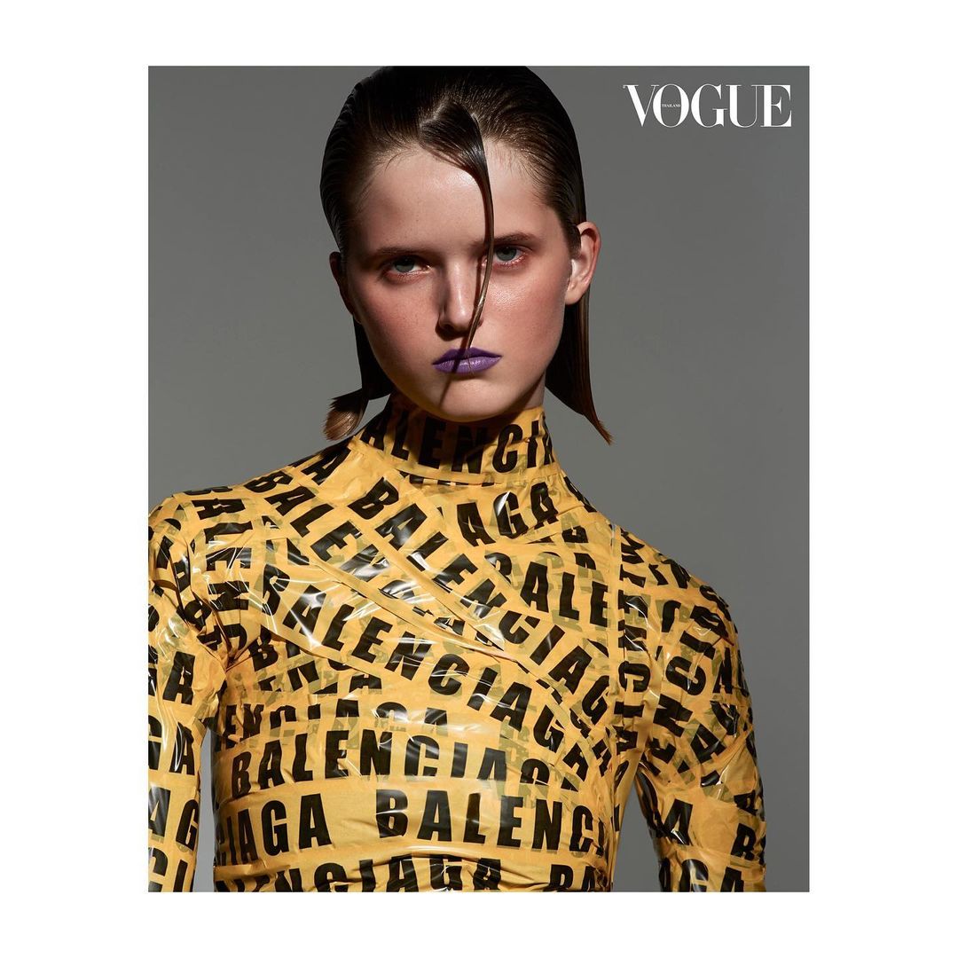 Close-up fashion shot of a young woman wearing a yellow long-sleeve top with black Balenciaga branding, against a solid gray backdrop.