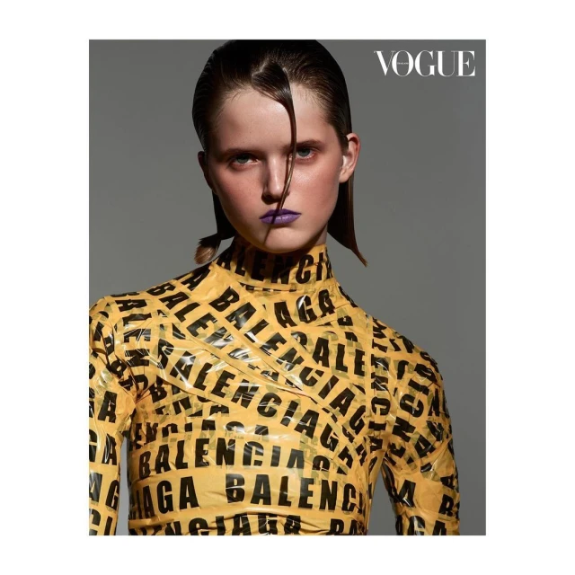 Close-up fashion shot of a young woman wearing a yellow long-sleeve top with black Balenciaga branding, against a solid gray backdrop.