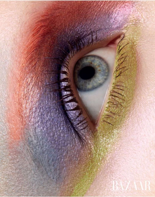 Close-up of a blue eye surrounded by orange, purple, and yellow eyeshadow with 'BAZAAR' logo at the bottom of the frame.