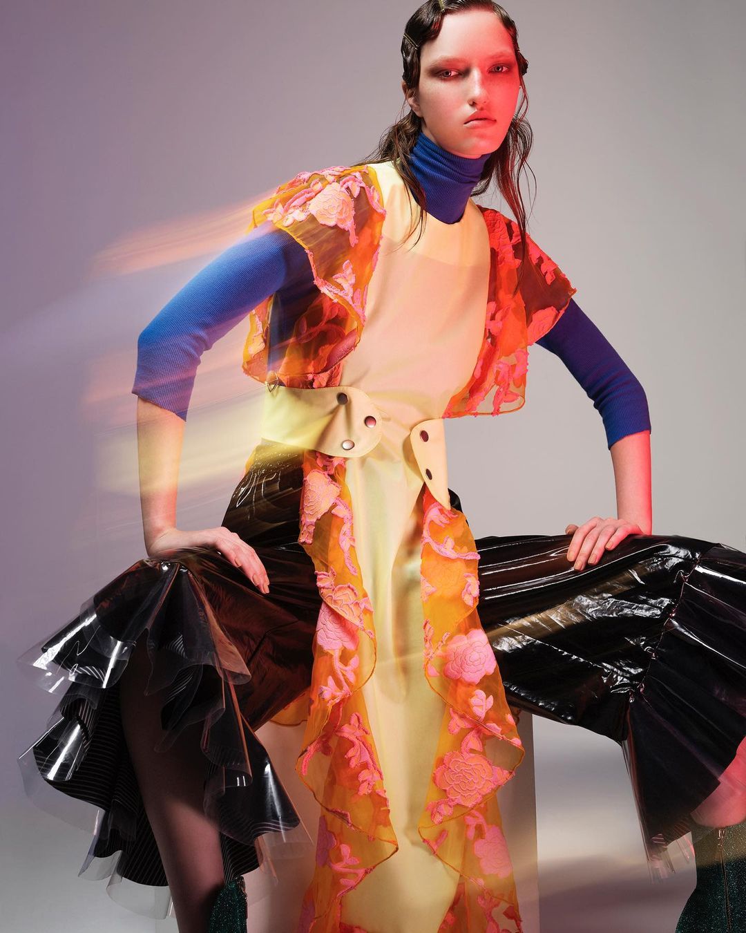 Fashion editorial shot of a female model in a yellow dress with orange floral accents and black vinyl pants in a photo studio.