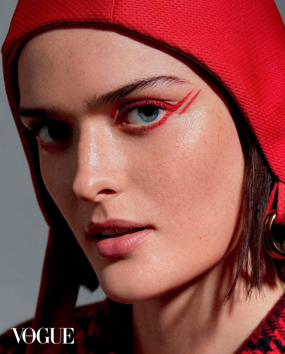 Close-up of a woman with red eye makeup and a red head covering, gazing directly at the camera. Beauty shot against a grey backdrop.
