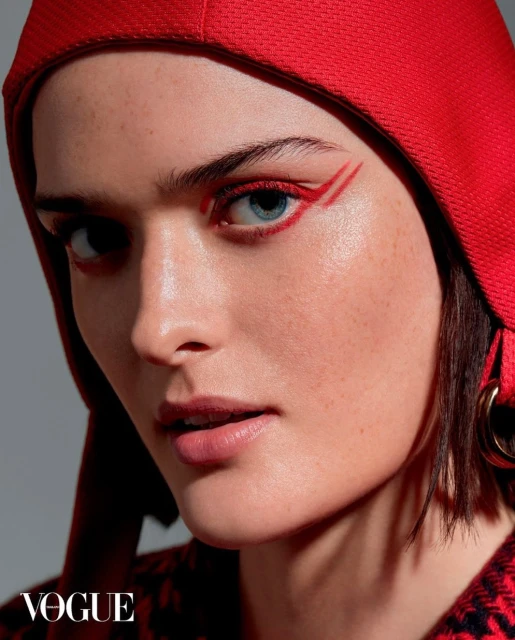 Close-up of a woman with red eye makeup and a red head covering, gazing directly at the camera. Beauty shot against a grey backdrop.